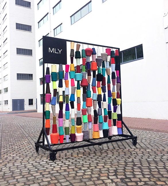 Installatie met gekleurde garenklossen op een verrijdbaar frame met MLY-logo
